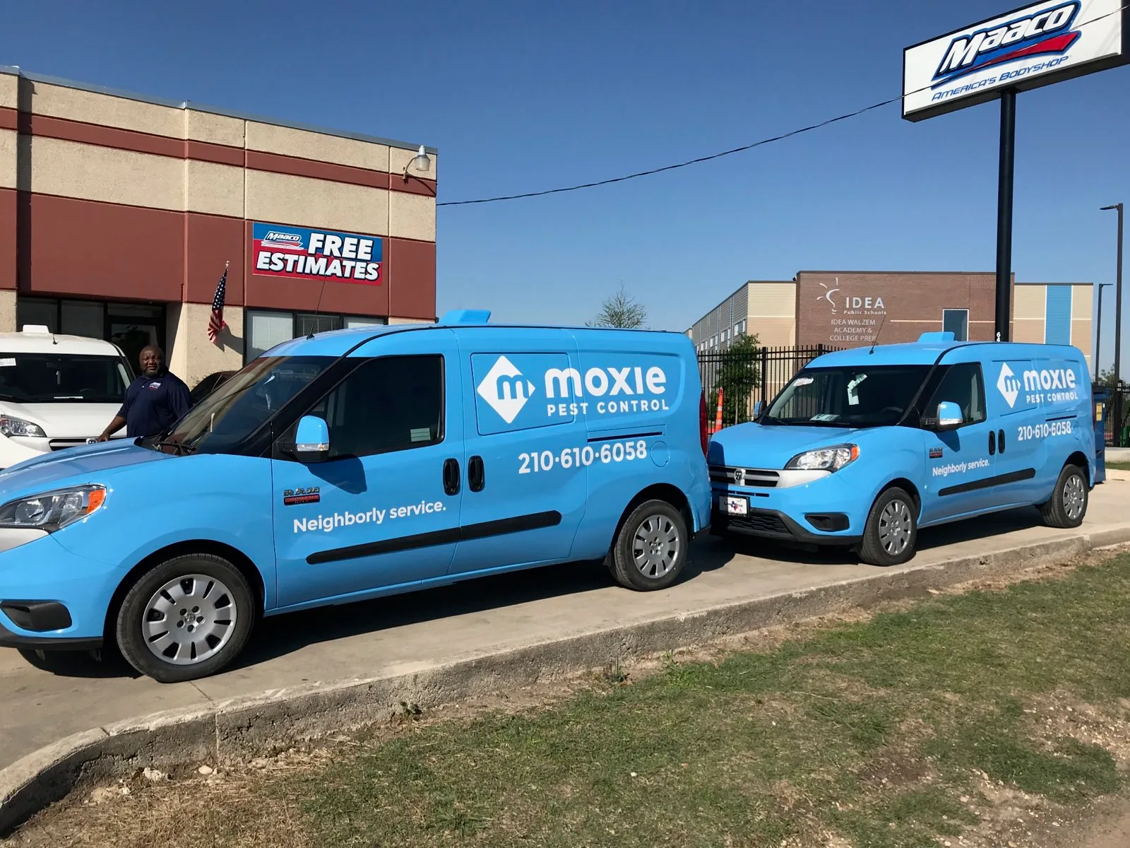 Two blue branded cargo vans parked in front of an auto body shop with a person standing beside one of them.