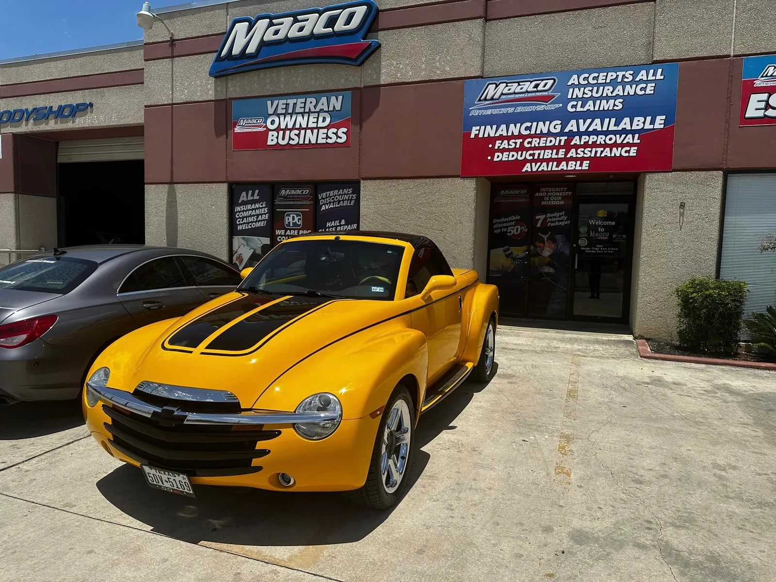 A yellow convertible pickup with black hood stripes parked on pavement outside a body shop storefront, with a silver sedan beside it.