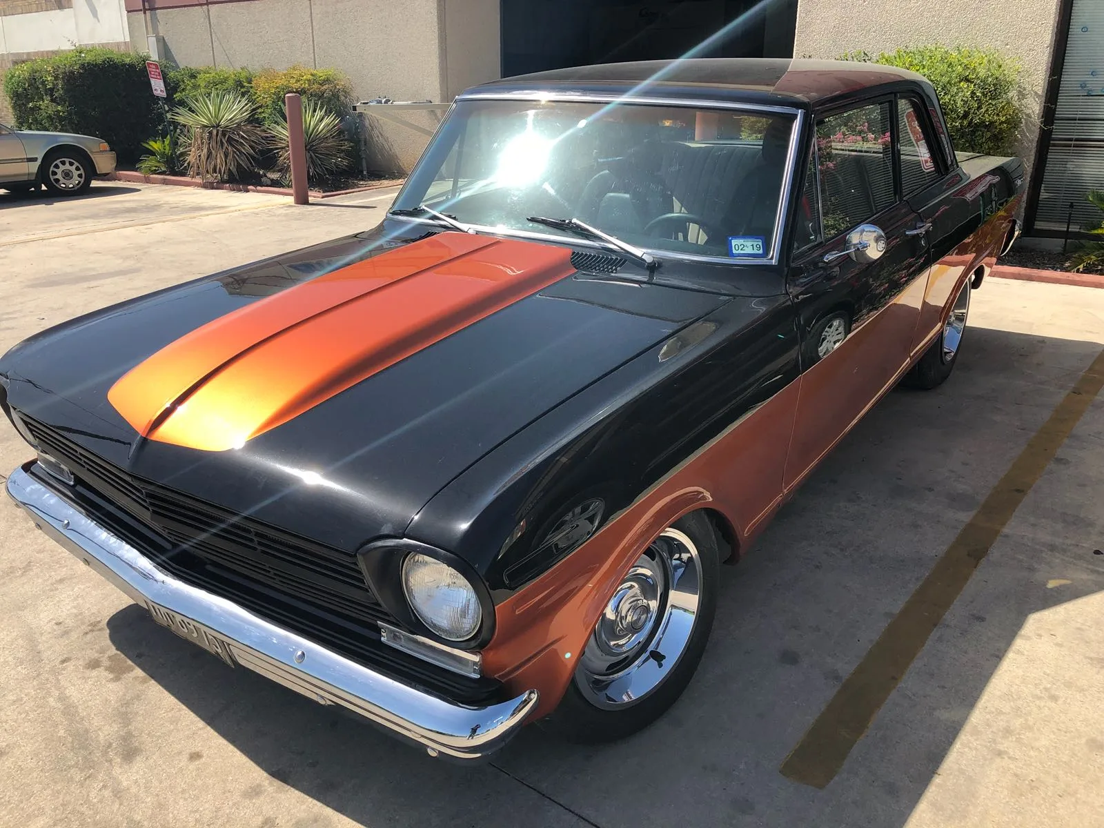A classic black two-door car with an orange hood stripe and chrome wheels parked in a lot.